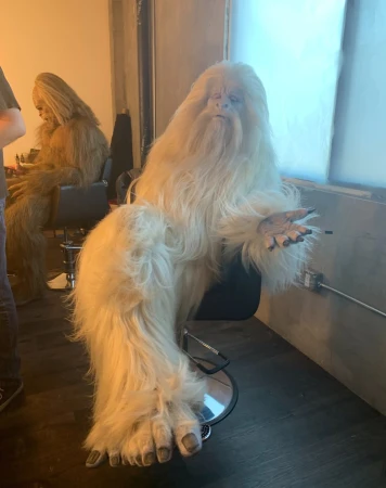 Person in a yeti costume sitting on a chair with another yeti costume in the background in an indoor setting.